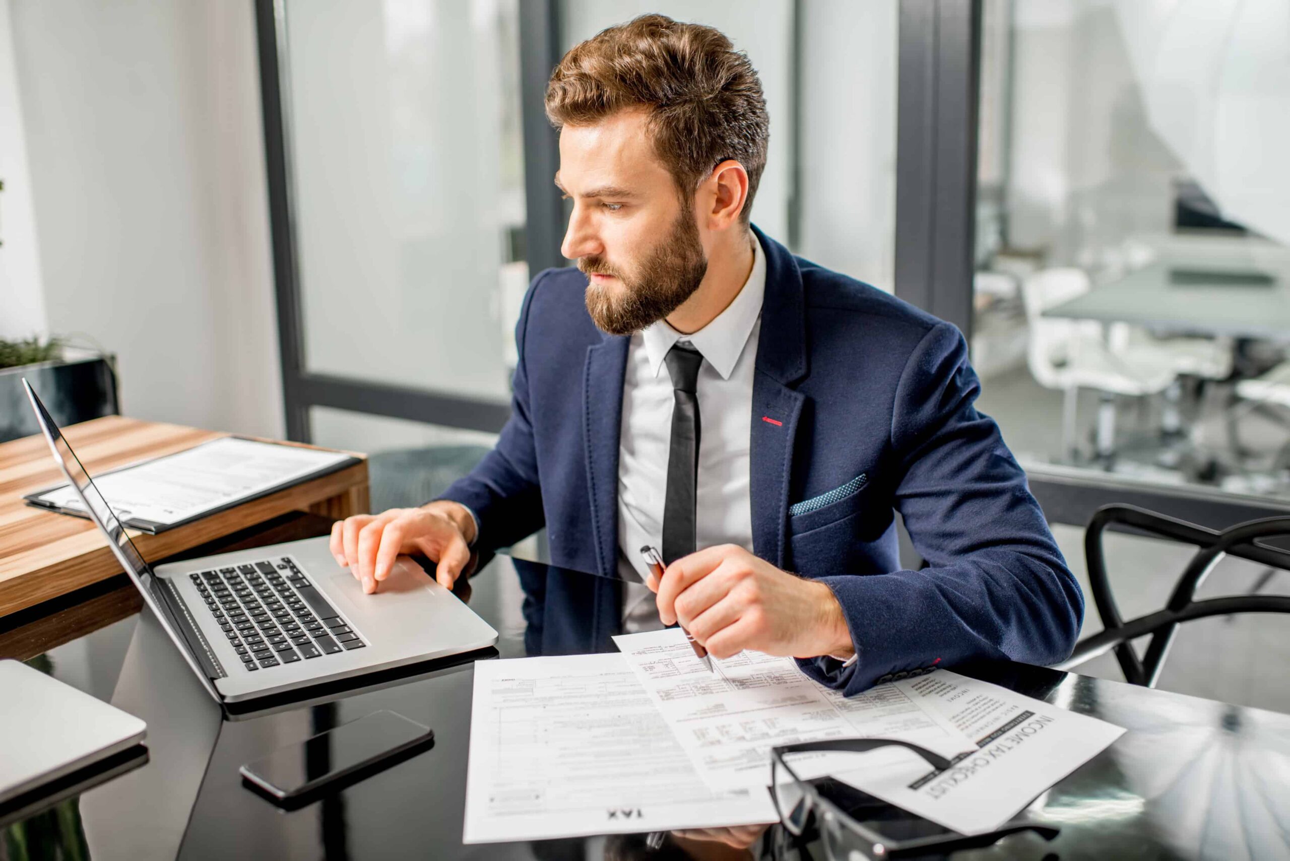 A man in a suit sits at a desk using a laptop, holding a pen and looking at documents labeled "TAX" with a smartphone and eyeglasses nearby in a modern office setting.